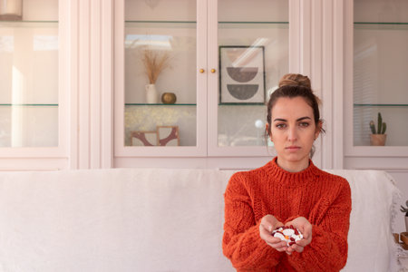 Young woman sitting in living room and demonstrating handful of different pills while looking at cameraの写真素材