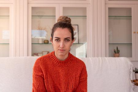 Young woman with dark hair wearing warm orange sweater sitting on white sofa at home looking at cameraの写真素材