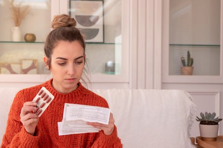 Focused young lady sitting on sofa with medications and reading drug prescription after visiting hospitalの写真素材