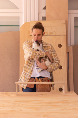 Handyman with adorable cat creating shelf. Male carpenter in tool apron standing near workbench with cute cat against boards and wooden detailsの写真素材