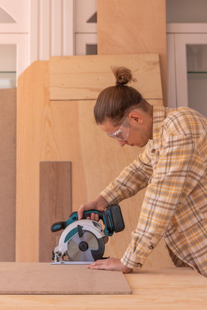 Handyman with circular saw working with wood. Side view of male carpenter in goggles cutting wooden board with circular saw on workbenchの写真素材