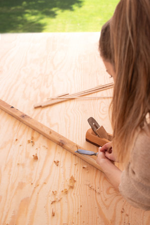 Craftswoman carving wood with chisel. Concentrated female joiner in apron gouging wood with chisel near jack planeの写真素材