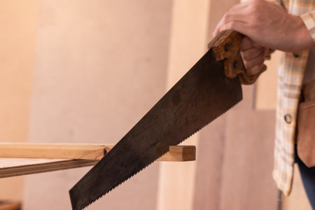 Crop male carpenter cutting wooden detail with saw while standing at workbench and working in joinery workshopの写真素材