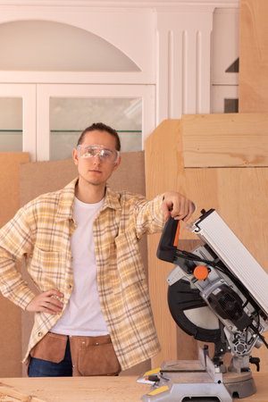 Young male carpenter with standing in home carpentry shop against wood planks near modern mitre sawの写真素材