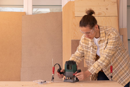 Young concentrated workman wearing checkered shirt while working with router machine in home workshopの写真素材