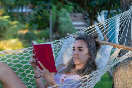 Young beautiful woman sitting in a hammock in summer concentrated reading a red book with natural environment in backgroundの写真素材