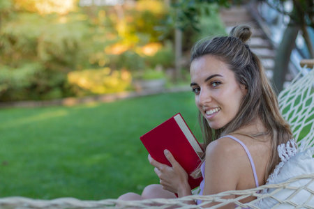 Happy smiling woman sitting in a hammock looking to camera while holding a book and copy space in the garden in summerの写真素材