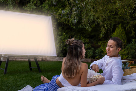 Happy young couple looking at each other and watching a movie in the outdoor summer cinema with a bowl of popcorn in the gardenの写真素材