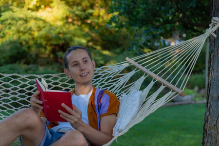 Young man sitting relaxed in a hammock reading a red book and looking to the side enjoying the natural environment with fresh airの写真素材
