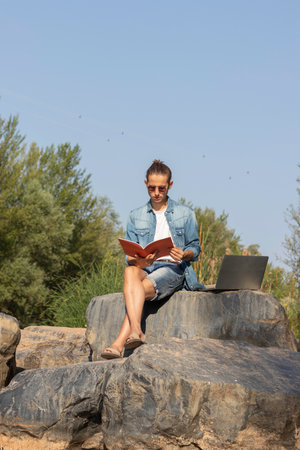Low angle portrait of a university student reading a magazine next to his laptop into de woods with copy spaceの写真素材