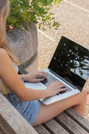 Vertical back view of unrecognizable young woman working as a freelancer on a laptop computer sitting on a park bench in summerの写真素材