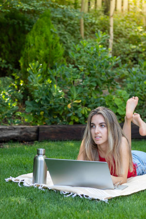 Vertical view of young beautiful woman looking to the side while using her laptop for her remote job in the parkの写真素材