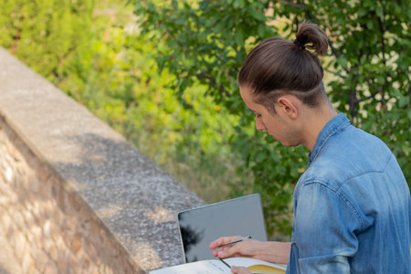 Young man with bun hairstyle looking down to his notebook enjoying leisure activity in the nature with laptop computerの写真素材