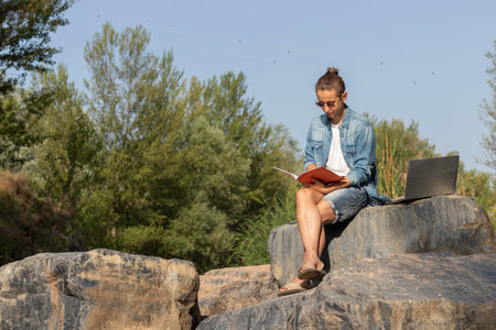 Young man enjoying remote employment focused on reading a catalog by his laptop in the woods in springの写真素材