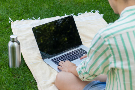 Bakc view of unrecognizable young online using a laptop on a towel and a bottle of water on the side in the gardenの写真素材