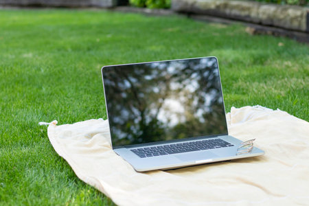 Isolated laptop on a towel in the garden reflecting the tree leaves in the screen with eyeglasses on top of it in spring dayの写真素材