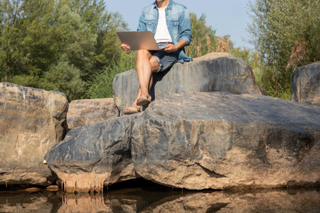 Unrecognizable portrait of a young man working with his laptop sitting on a red one next to the bed of a river in Spainの写真素材