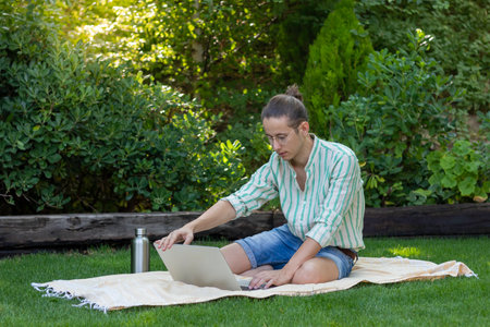 Young man with bun hairstyle and eyeglasses on opening his laptop to start working on his first online job sitting in the gardenの写真素材