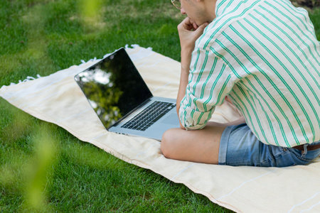 Back view of faceless young man concentrated using the computer sitting outdoors under a tree in springの写真素材