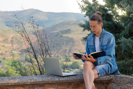Concentrated young digital nomad drawing on his notebook with a laptop reading on the floor with a landscape in the backgroundの写真素材