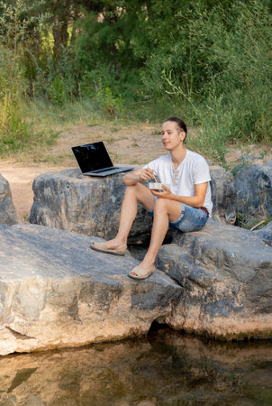 Digital nomad young male taking advantage of his break to eat a picnic in nature next to a green riverの写真素材