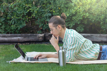 Side view of young man working relaxed wearing bun hairstyle and eyeglasses leaning on the grass in nature environmentの写真素材