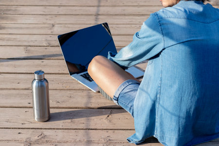 Back view of unrecognizable young man sitting on wooden floor working online on laptop as digital nomad in springの写真素材