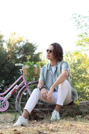 Vertical view of young man sitting on a tree trunk in the park relaxing with his pink vintage bicycle in springの写真素材