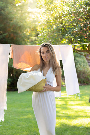 Vertical view of young gergeous woman standing up in front of the hanging clothes looking to camera in the garden with a basket in handsの写真素材