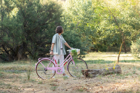 Side view of unrecognizable back looking young long haired man holding a pink vintage cute bike in the city parkの写真素材