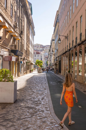 Vertical View of a Young Woman in an orange Dress Walking Down a Cobblestone Street in Lisbon, Portugal, Surrounded by Historic Buildings,の写真素材