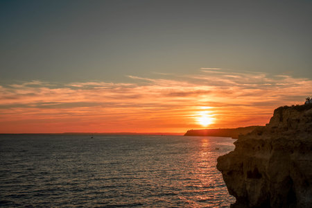 A Tranquil Sunset of a Rocky Cliff Overlooking the Ocean, Capturing the Warm Glow of the Setting Sun Against the Deep Orange Skyの写真素材