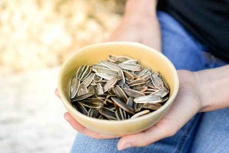 Closeup view of unrecognizable woman in black holding plenty of sunflower seeds in a brown bowl in backgroundの写真素材
