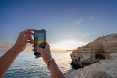 Golden Hour Moment: Young Man Hands Capturing the Ocean Sunset of beautiful cliff shoreの写真素材