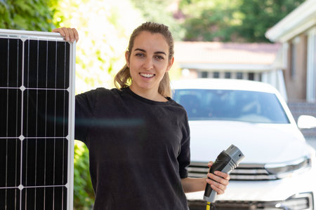 Portrait of young woman smiling holding a solar panel showing the source of energy for her electric white car and a plug on handの写真素材