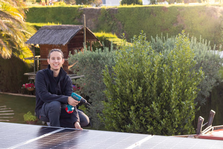 Young woman technician working in the installation of solar panels on the roof of a house with electric screwdriver on hand surrounded by vegetationの写真素材
