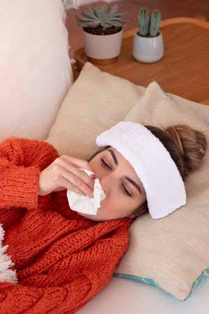 From above of ill female with influenza and high temperature blowing running nose while lying on comfortable sofa during sick day at homeの写真素材