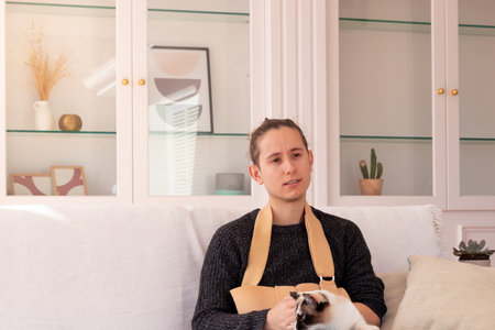 Young male in casual clothes with broken hand wearing arm sling looking away while sitting on sofa with cat in living room at homeの写真素材