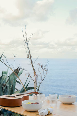 Wooden table with acoustic guitar bowls of muesli cups spoons and folded towels set near green coastal plant on seashore against waving seaの写真素材