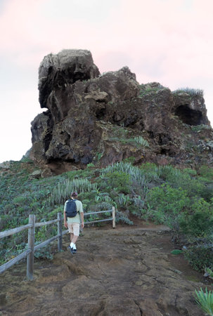 solitary male hiker, in casual attire, ventures towards a towering rock formation amidst lush greenery under the soft hues of the evening skyの写真素材