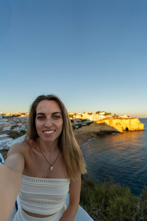 Vertical selfie of Young long blonde hair Woman in a White Dress Overlooking a Coastal Townの写真素材