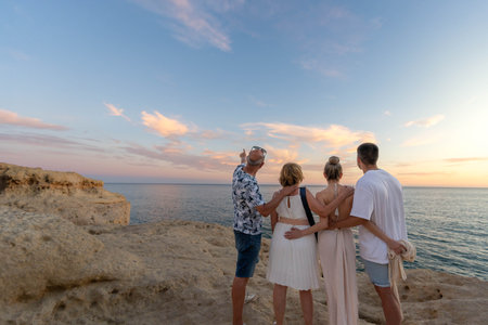 Back view of man pointing at sky while embracing with family standing on rocky cliff admiring picturesque sunset over ocean during summer tripの写真素材