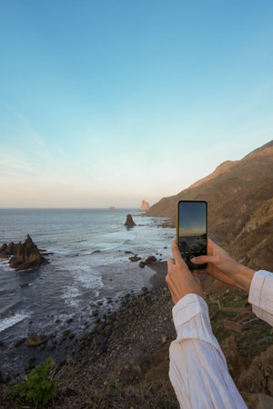 A close-up of hands holding a smartphone, capturing the serene beauty of a rocky coastal landscape bathed in the warm hues of the golden hourの写真素材