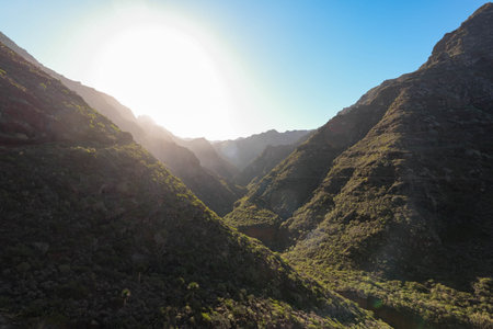 Breathtaking horizontal shot captures the serene moment of sunrise over a rugged mountain range. The steep slopes, covered with green vegetation, are softly illuminated by the dawn light, creating a tranquil atmosphere under the clear, blue skyの写真素材
