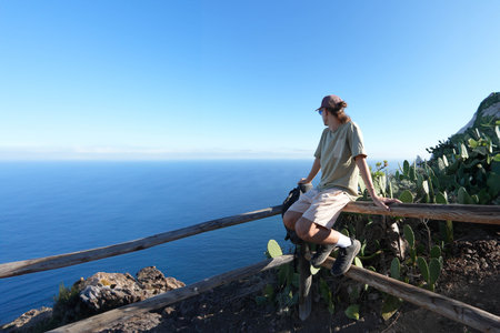 solo traveler, in casual hiking attire, sitting on a wooden railing at the edge of a mountain. turned towards the vast ocean, suggesting a moment of serene contemplation.の写真素材