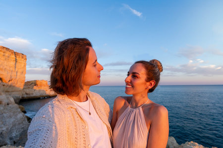 Young man embracing smiling girlfriend while standing on rocky cliff against blue sea and sundown sky during romantic tripの写真素材
