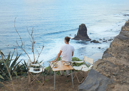 Young man immersed in the serene beauty of a rocky shoreline while sitting on the edge of a camping picnic table with foodの写真素材