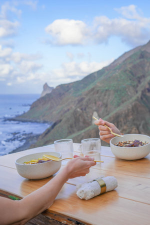 An intimate outdoor dining scene, hands serving food, with a breathtaking ocean and mountain backdrop in daylightの写真素材