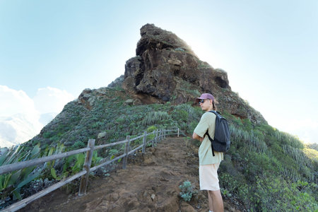 solo hiker from the rear, standing on a dirt path in Tenerife. Dressed in casual hiking attire and carrying a backpack, the individual is seen admiring a majestic mountain peak covered in lush greenery.の写真素材