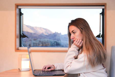 A digital nomad is focused on a laptop while working from the comfort of her van, showcasing the freedom and lifestyle of remote work against a backdrop of tranquil scenery at dusk.の写真素材